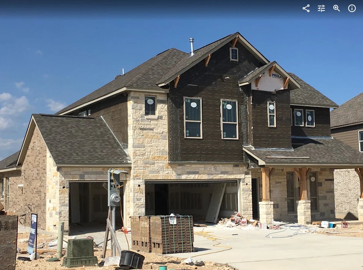 Two-story house under construction with stone and dark siding, open garage, and construction materials on the front lawn