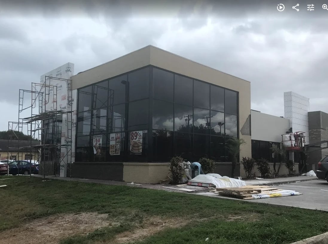 Modern industrial building with large black windows, tan exterior panels, and scaffolding on the left side under cloudy sky