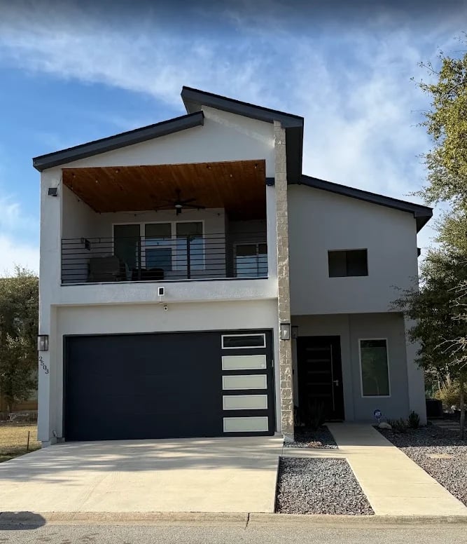 Modern two-story house with white and gray exterior, black garage door, front balcony, and metal railing under partly cloudy sky